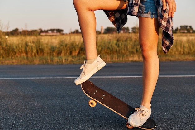 Pessoa desconhecida do sexo feminino com pernas longas e bonitas usando tênis branco andando de skate, patinador sem rosto andando de skate no verão, tiro ao ar livre.