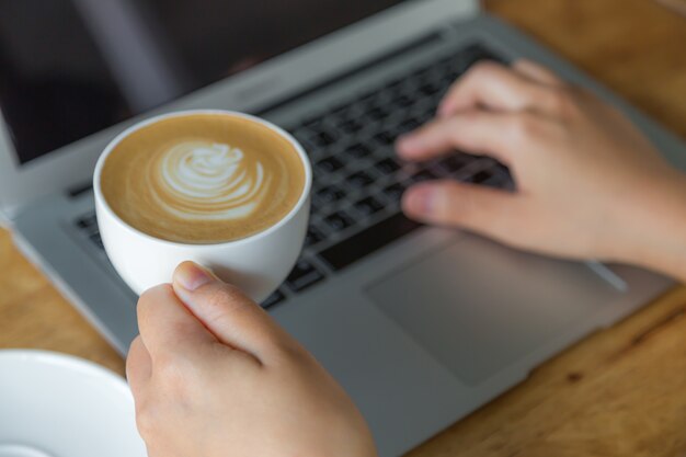 Pessoa digitando em um laptop segurando uma xícara de café