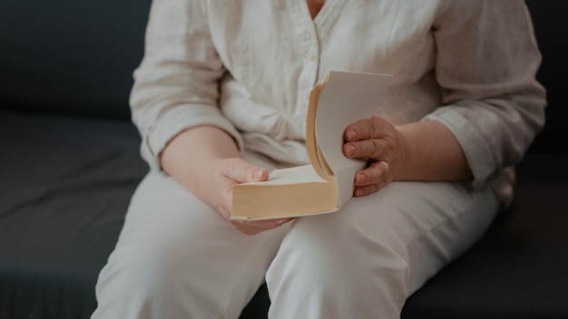 Pessoa idosa segurando livro de ficção para ler e relaxar no sofá. mulher sênior lendo história de literatura em papel, aproveitando a aposentadoria em casa. pensionista inteligente com foco no conhecimento. fechar-se