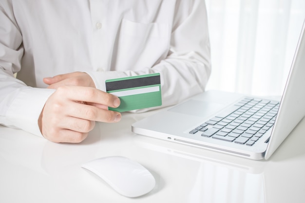 Foto grátis pessoa segurando um cartão de crédito verde com um laptop e um mouse de computador em uma mesa branca