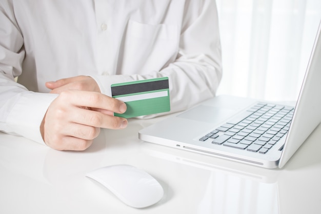 Pessoa segurando um cartão de crédito verde com um laptop e um mouse de computador em uma mesa branca