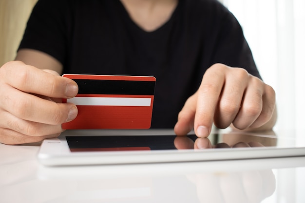 Pessoa segurando um cartão de crédito vermelho sobre um tablet em uma superfície branca