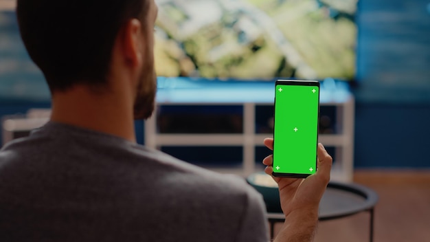 Pessoa segurando um telefone celular com tela verde verticalmente no visor, sentado na sala de estar em casa. Fundo isolado com chroma key e maquete para modelo de mídia no espaço da cópia