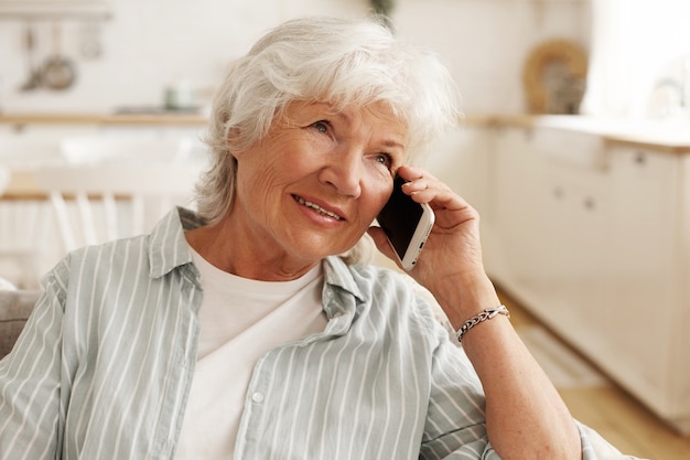 Pessoas, aparelhos eletrônicos modernos, tecnologia e comunicação. Mulher idosa idosa com cabelo curto e grisalho, desfrutando de uma bela conversa ao telefone, sentada no sofá, segurando o celular na orelha e sorrindo