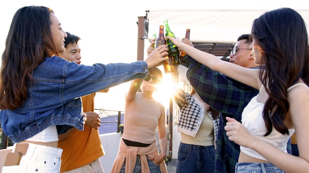 Pessoas bebendo álcool na festa no terraço do pôr do sol amigos asiáticos tinindo com garrafas de cerveja no terraço