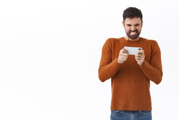 Pessoas de tecnologia e conceito de comunicação retrato de homem caucasiano bonito animado tentando passar de nível difícil aperta os dentes em pé intenso e toca na tela do celular como jogo de tiro