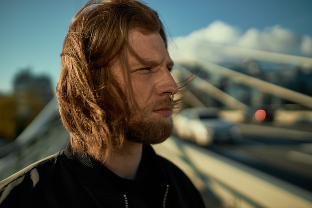 Pessoas e conceito de estilo de vida da cidade. Retrato de um cara barbudo elegante e atraente com cabelo ruivo bagunçado e piercing no nariz contrastando com o fundo da paisagem urbana, com olhar sério e confiante