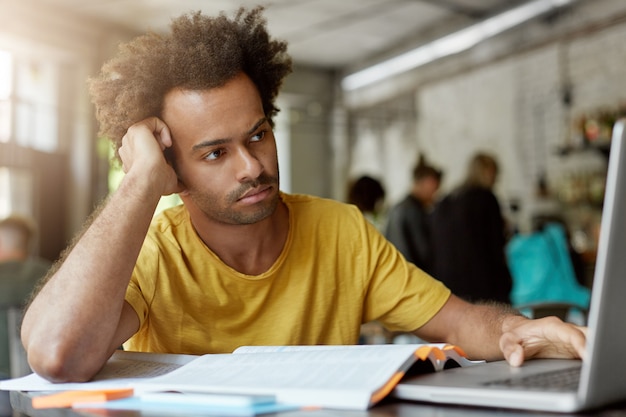 Pessoas, educação, tecnologia moderna e conceito de juventude. homem mestiço de pele escura com cabelo encaracolado sendo focado na tela do laptop com olhar pensativo inclinando-se para sua mão sentado em um café