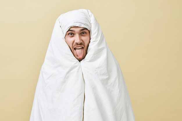 Pessoas, estilo de vida, conceito de hora de dormir. jovem emocional com a barba por fazer posando enrolado da cabeça aos pés em um cobertor, com preguiça de se levantar, mostrando a língua, provocando você