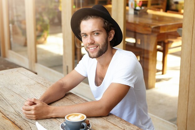 Pessoas, estilo de vida e lazer. Retrato interior de bonito jovem hippie elegante chapéu preto, sentado à mesa de café de madeira com caneca de cappuccino