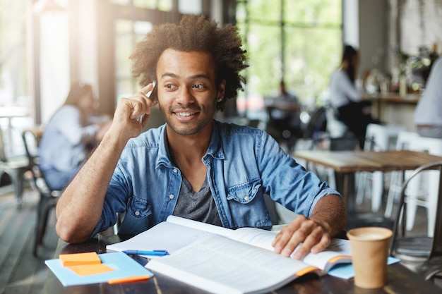 Pessoas, estilo de vida, educação e conceito de tecnologia moderna. foto sincera de um estudante afro-americano alegre com roupas elegantes, conversando no celular enquanto faz o dever de casa na cantina