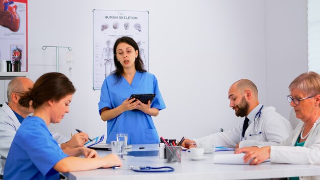 Pessoas médicas, reunião e planejamento com acionistas no escritório do hospital, sentado à mesa. Médicos e enfermeiras discutem ideias juntos, diagnósticos médicos e dados de apresentação usando um tablet