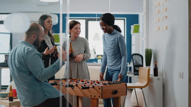 Pessoas multiétnicas curtindo o jogo na mesa de pebolim tomando bebidas alcoólicas no escritório depois do trabalho. homem e mulher jogando futebol a bordo enquanto colegas aproveitam a celebração.
