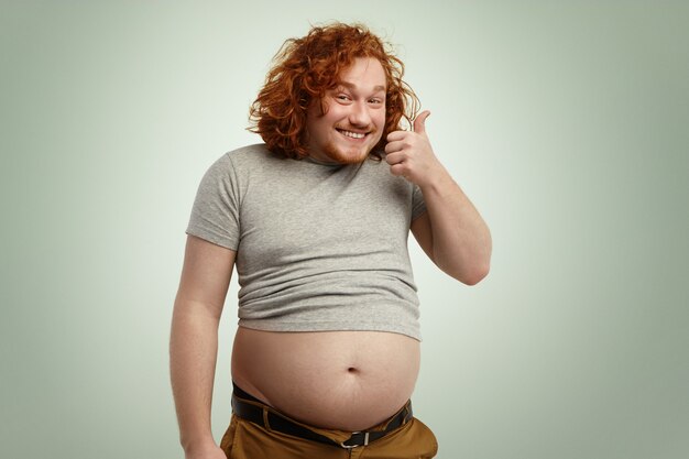Pessoas, nutrição, forma do corpo e conceito de estilo de vida saudável. Feliz jovem gordinho gordinho com cabelo ruivo cacheado e barba aparecendo, dizendo que ele está bem enquanto sua barriga está saindo