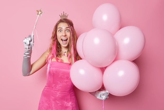 Foto grátis pessoas ocasião especial e conceito de entretenimento princesa jovem e bonita positiva usa coroa na cabeça vestido luvas longas faz seus desejos se tornarem realidade segura varinha mágica bando de balões de hélio