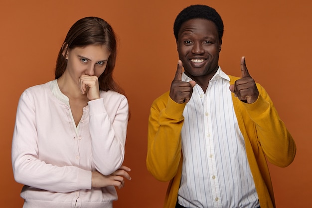 Foto grátis pessoas, raça e etnia. jovem afro-americano animado e feliz de bom humor, sorrindo alegremente, apontando os dedos na frente, uma linda garota europeia de cabelos compridos segurando o punho contra a boca, rindo