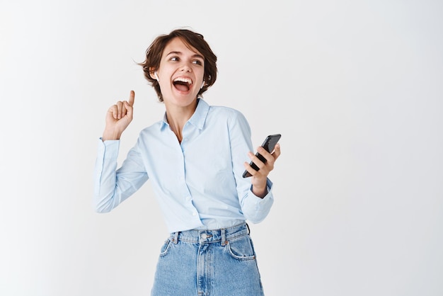 Pessoas reais e conceito de tecnologia Mulher caucasiana feliz dançando com smartphone na mão rindo e sorrindo ouvindo música em fones de ouvido sem fio fundo branco