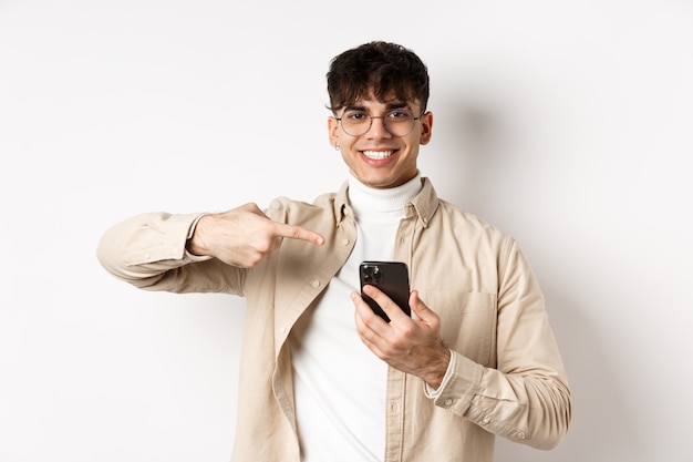 Pessoas reais. Jovem bonito de óculos apontando o dedo para a tela do smartphone, mostrando uma promoção online, em pé sobre um fundo branco