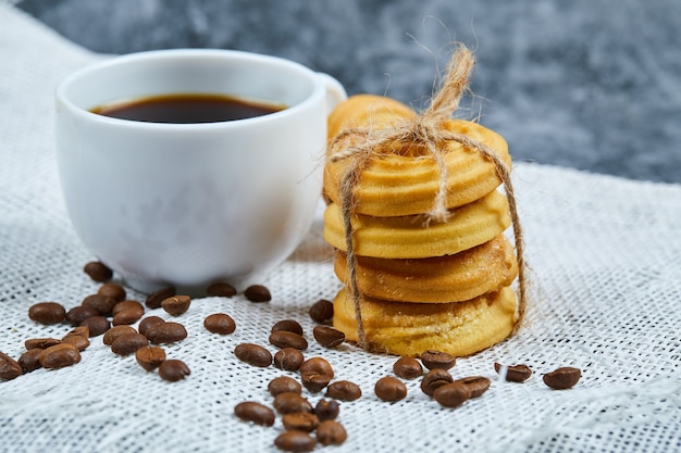 Pilha de biscoitos com grãos de café e uma xícara de café em uma toalha de mesa branca.