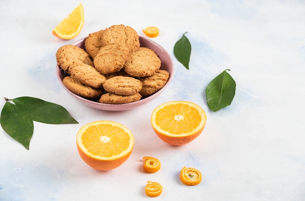 Pilha de biscoitos em uma tigela rosa e meio corte de laranjas com folhas sobre a mesa branca.