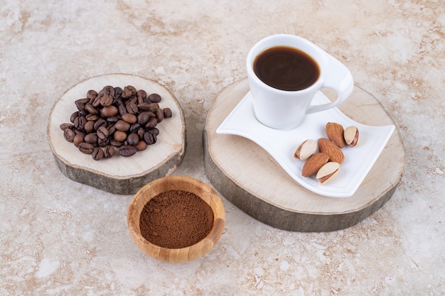 Pilha de grãos de café em uma placa de madeira ao lado de uma pequena tigela de café moído e uma xícara de café com amêndoas e pistache