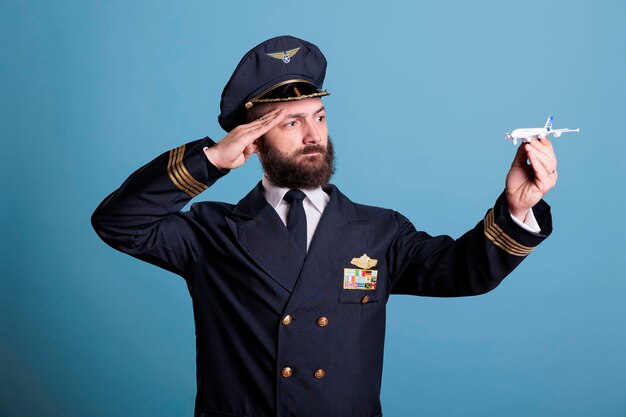 Piloto de uniforme saudando o modelo de avião pequeno, capitão de avião brincando com brinquedo de jato comercial de passageiros. Grave membro da tripulação de aeronaves com distintivo de asas na jaqueta, tiro médio de estúdio