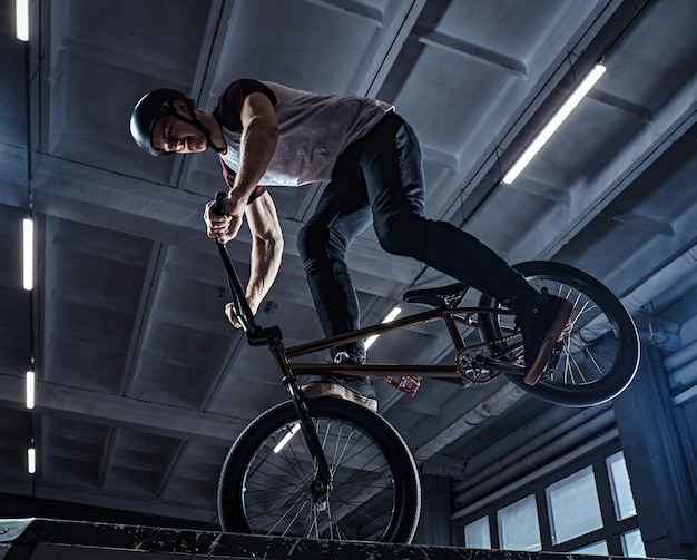 Piloto profissional de BMX em capacete protetor realizando truques no skatepark dentro de casa