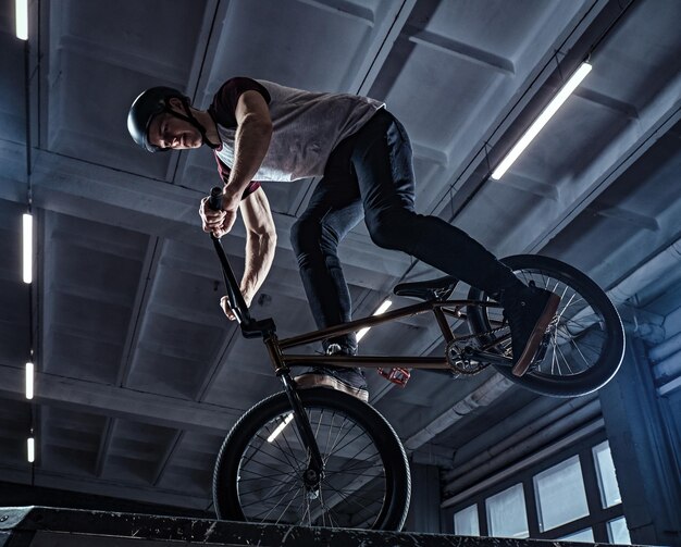 Piloto profissional de BMX em capacete protetor realizando truques no skatepark dentro de casa