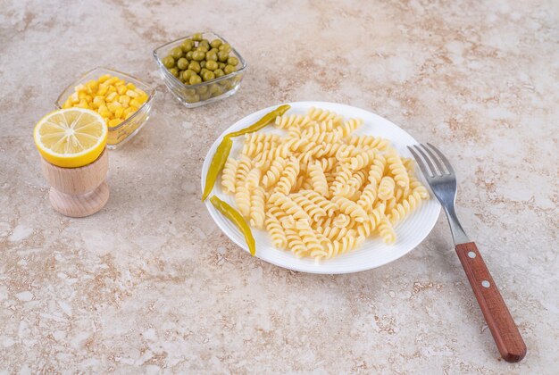 Pimentas em conserva decorando macarrão cozido, com tigelas de ervilhas, milho e um limão fatiado em superfície de mármore.