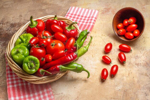 Pimentas verdes e vermelhas, de frente, pimentas, tomates em uma cesta de vime, tomates cereja espalhados da tigela, toalha de cozinha em fundo âmbar