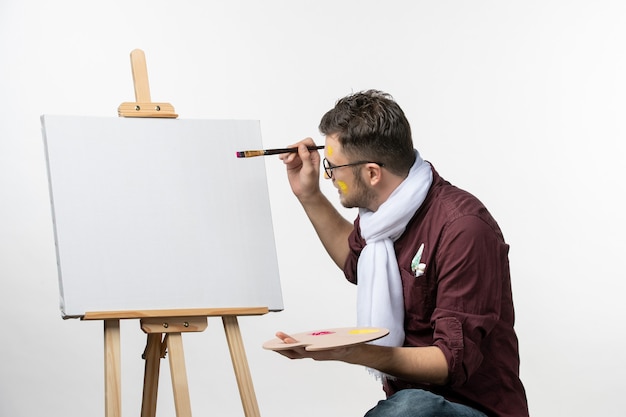 Foto grátis pintor de frente para o homem desenhando no cavalete com pincel e tintas na parede branca