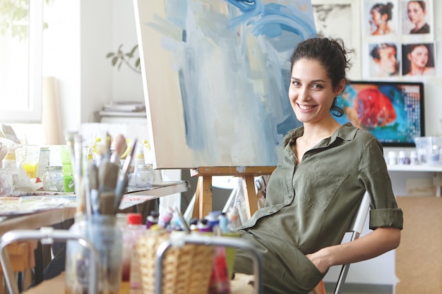 Pintor profissional feminino, sentado na cadeira no estúdio de arte, mantendo as mãos nos bolsos da camisa dela, sorrindo suavemente enquanto descansava depois de desenhar imagens com aquarelas. pessoas, passatempo, conceito de pintura