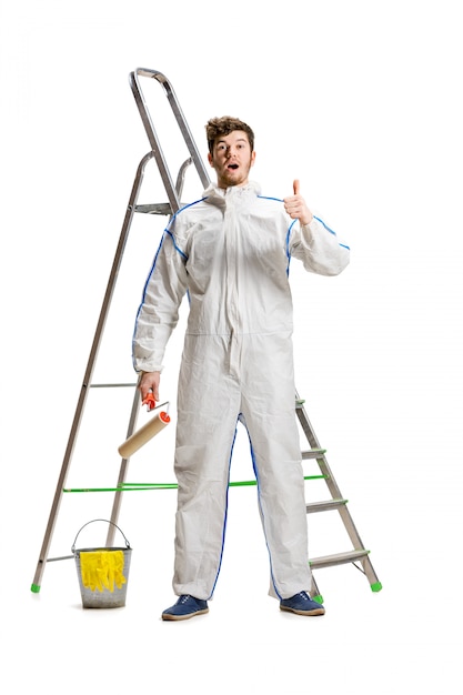 Pintura masculina nova do decorador com um rolo de pintura e uma escada isolada na parede branca.