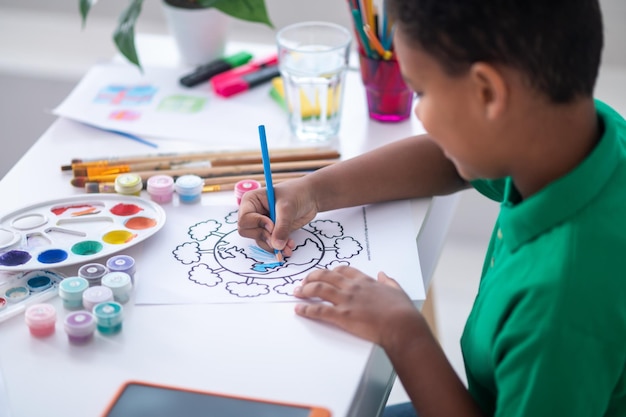 Pintura. Menino em idade escolar de pele escura desenhando com lápis azul de lado para a câmera enquanto está sentado à mesa na sala sob a luz do dia