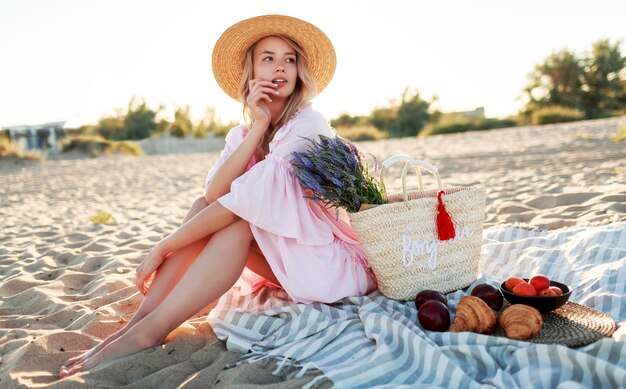 Piquenique na zona rural perto do oceano. Mulher jovem graciosa com cabelos loiros ondulados no elegante vestido rosa, curtindo as férias e comendo frutas.