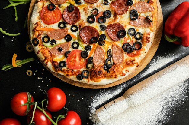 Pizza de salame vista superior com tomate pimentão e azeitonas em uma bandeja com farinha e rolo