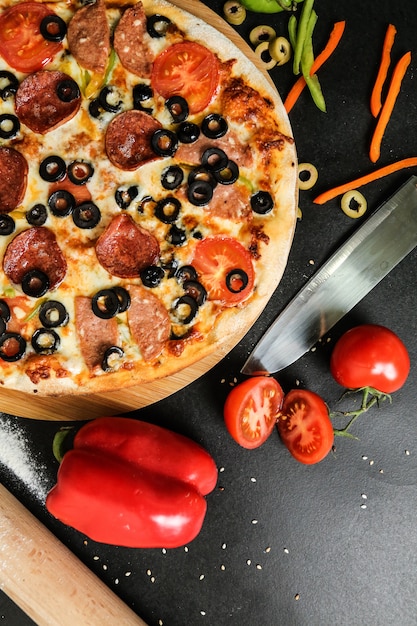 Foto grátis pizza de salame vista superior em stand com faca tomate azeitonas e pimentão na mesa preta