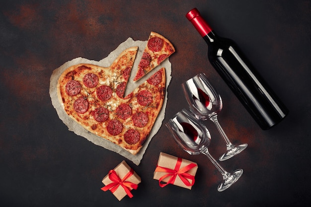 Pizza em forma de coração com mussarela, lingüiça, garrafa de vinho e dois copo de vinho. cartão de dia dos namorados em fundo enferrujado.