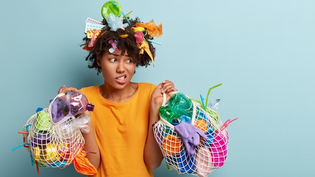 Plano horizontal de descontentamento feminino afro-americano de pele escura franze o rosto enquanto recolhe o lixo, limpa o ambiente, segura sacos de plástico com rede, desvia o olhar e pede para não jogar lixo na natureza