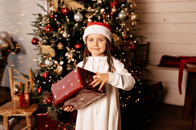 Época de Natal, criança alegre com um chapéu de Natal e um presente de Natal posando sobre uma árvore de Natal