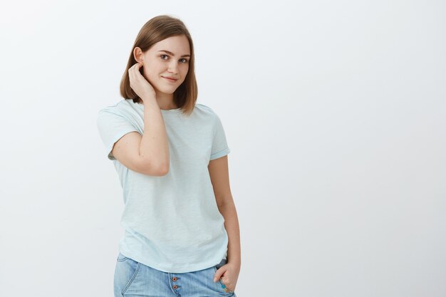 Podemos conversar um pouco. Tímida, fofa e gentil jovem morena atraente com corte de cabelo curto, passando o cabelo atrás da orelha e olhando com ternura com um leve sorriso simpático posando sobre uma parede cinza