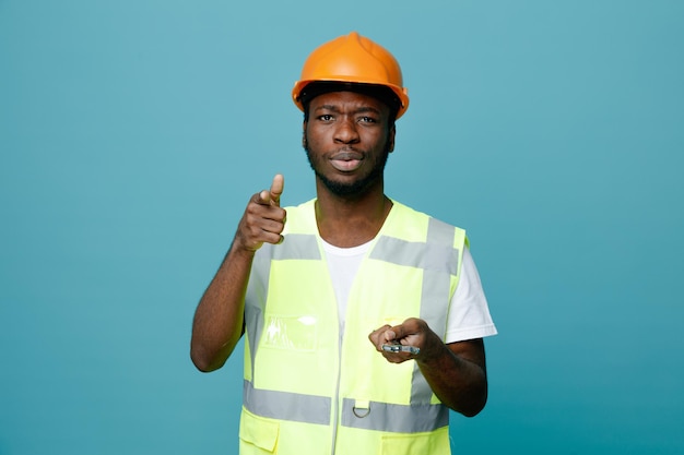 Pontos confiantes na câmera jovem construtor americano africano de uniforme segurando a chave aberta isolada em fundo azul