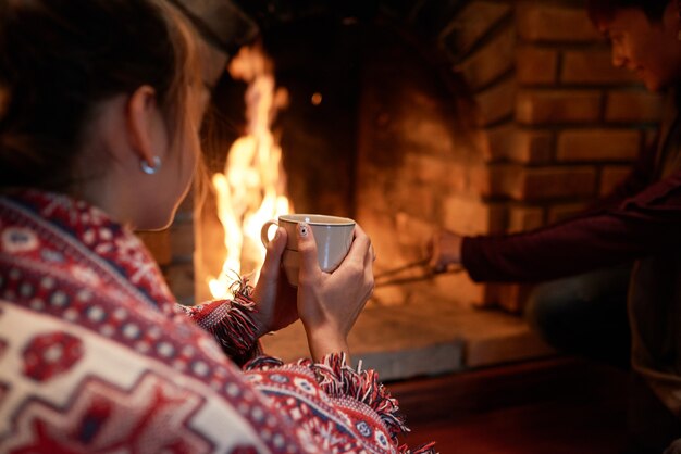 Por cima da foto do ombro da mulher aquecendo as mãos na caneca de chá quente sentada à lareira, o namorado lidando com carvão