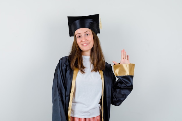 Pós-graduação feminino acenando com a mão para saudação em vestido acadêmico e olhando alegre. vista frontal.