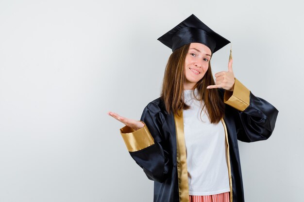 Pós-graduação feminino mostrando gesto de telefone em vestido acadêmico e olhando feliz. vista frontal.