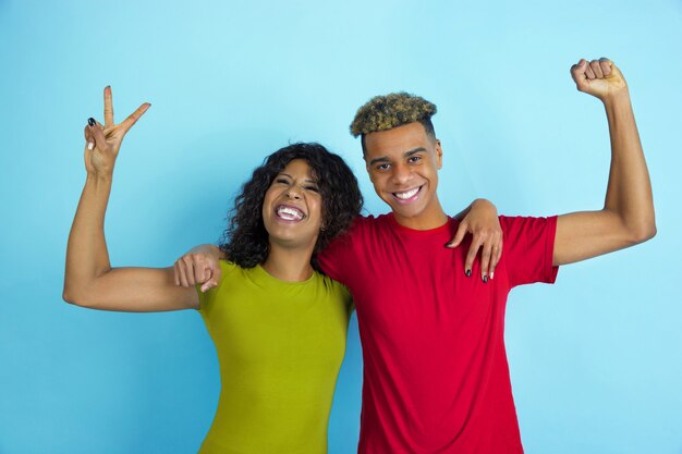 Posando como vencedores, rindo. Jovem emocional afro-americano e mulher com roupas coloridas na parede azul.