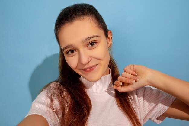 Posando para tirar uma selfie. Retrato de uma menina adolescente caucasiana sobre fundo azul. Bela modelo em moda casual. Conceito de emoções humanas, expressão facial, vendas, anúncio. Copyspace. Parece feliz.