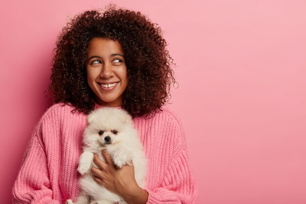 Positiva adolescente de pele escura com penteado afro espesso, posa com spitz branco no estúdio rosa, pensa em fazer um piquenique na natureza.