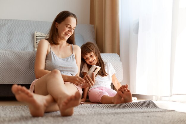 Positiva feliz atraente jovem adulta sentada no chão com sua filha com um telefone inteligente nas mãos, sorrindo, expressando emoções otimistas, usando o celular com a criança.