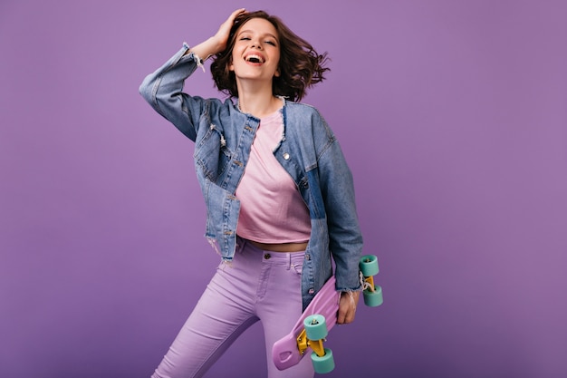 Foto grátis positiva garota branca em calças cor de rosa, posando com o skate. mulher encaracolada alegre sorrindo.